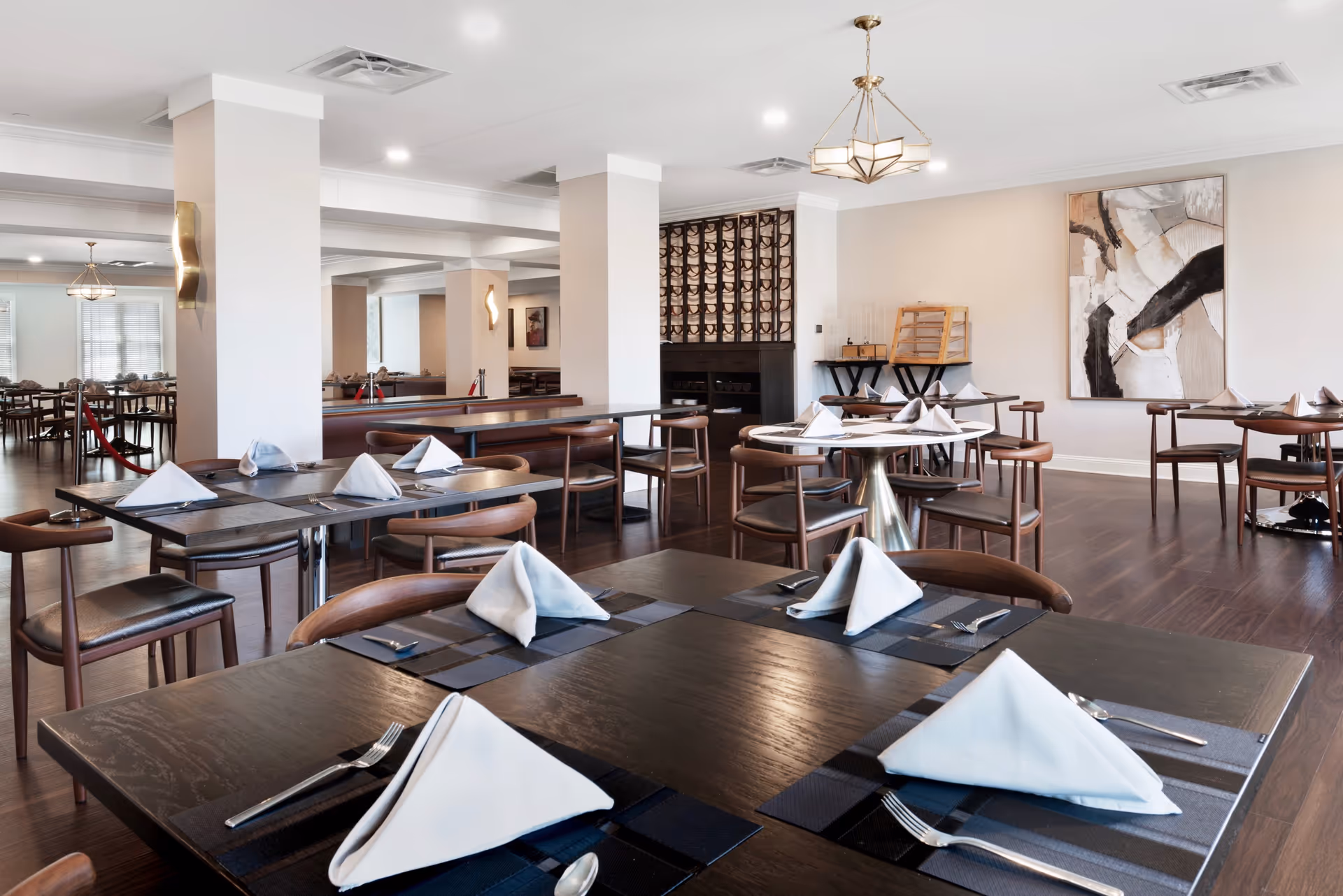 A spacious and modern dining room with multiple tables set with folded white napkins, silverware, and dark placemats. The room features wooden chairs, hardwood flooring, white walls, decorative lighting fixtures, and abstract artwork on the wall. There is a wine rack and a small buffet area in the background.