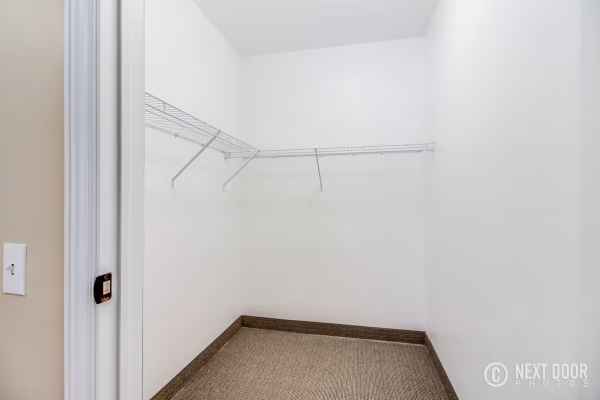 Empty walk-in closet with white walls, beige carpeted floor, and white wire shelving on two walls.