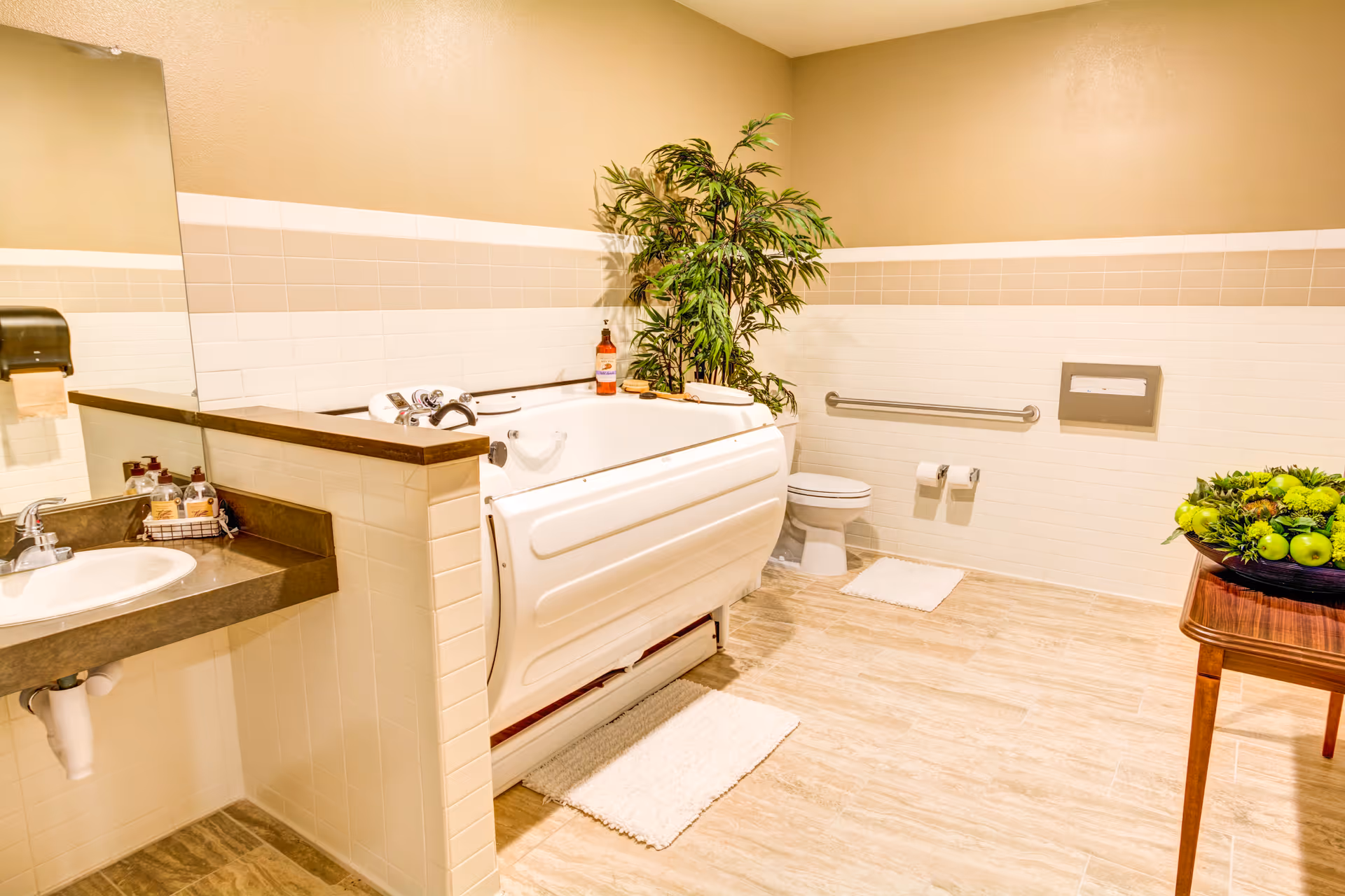 A spacious bathroom with a large white bathtub equipped with safety handles, a toilet with grab bars, a sink with soap dispensers, a paper towel dispenser, and a decorative plant next to the bathtub. There is also a small table with a bowl of green apples and other greenery. The walls are tiled halfway up with beige and white tiles, and the floor has light-colored tiles with two white bath mats.