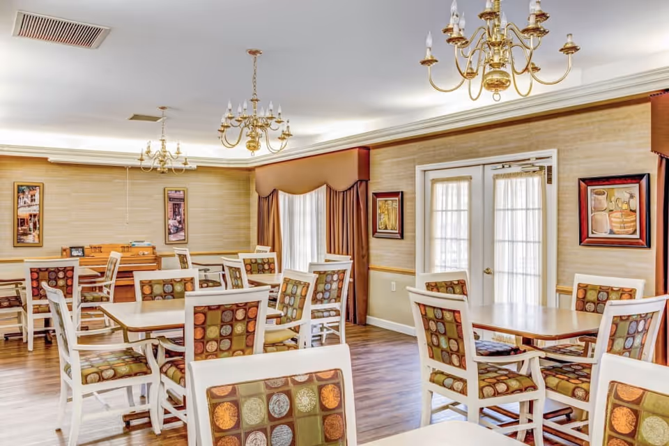 A bright dining room with multiple tables and chairs featuring patterned upholstery. The room has wooden flooring, beige walls with framed artwork, and large windows covered with curtains. Several gold chandeliers hang from the ceiling, and there is a piano against one wall.