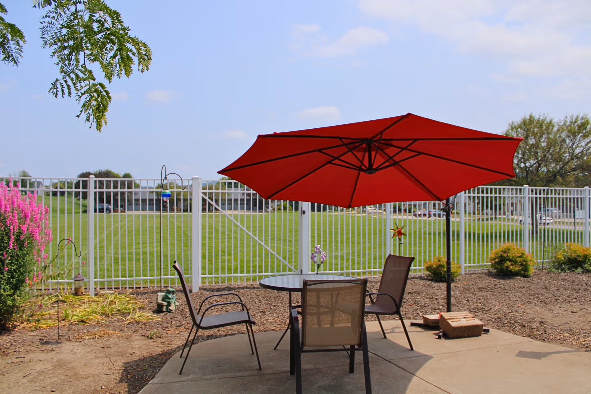 Patio seating area with a red umbrella and chairs overlooking a fenced grassy yard.