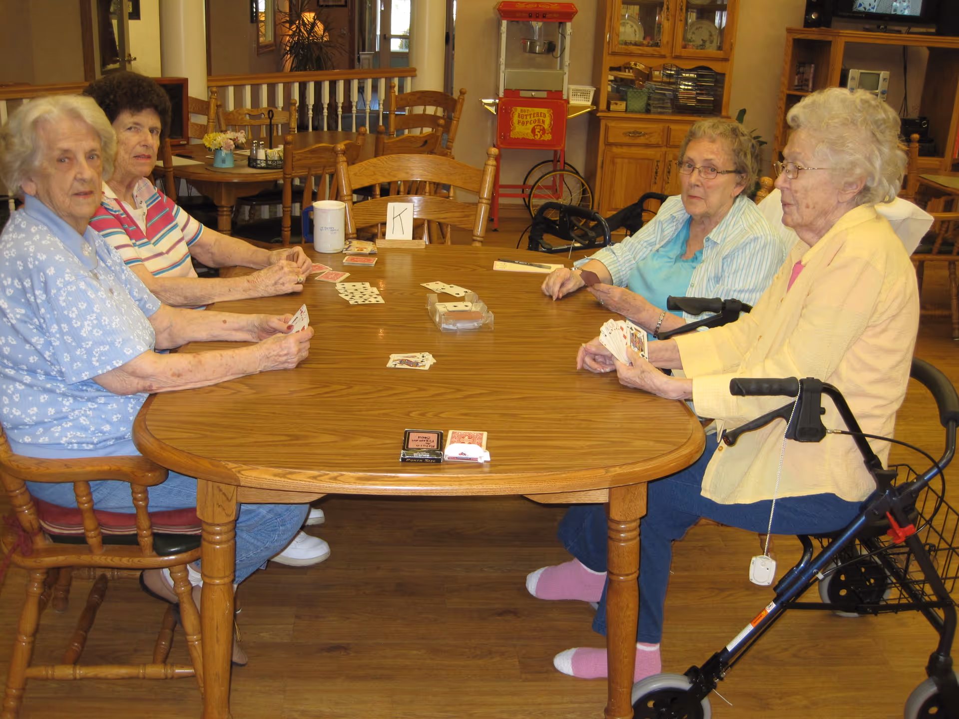 Four elderly women sitting around a wooden table playing cards in a communal room with wooden furniture and a popcorn machine in the background.