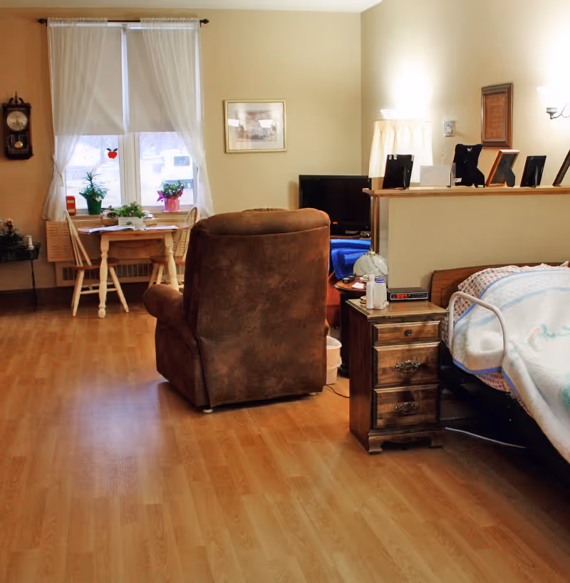 A cozy room with a brown recliner chair facing a TV on a stand. To the right is a bed with a colorful blanket and a wooden nightstand holding a lamp, clock, and some items. In the background, there is a small wooden dining table with two chairs near a window with white curtains and potted plants on the windowsill. The room has wooden flooring and beige walls with framed pictures and a wall clock.