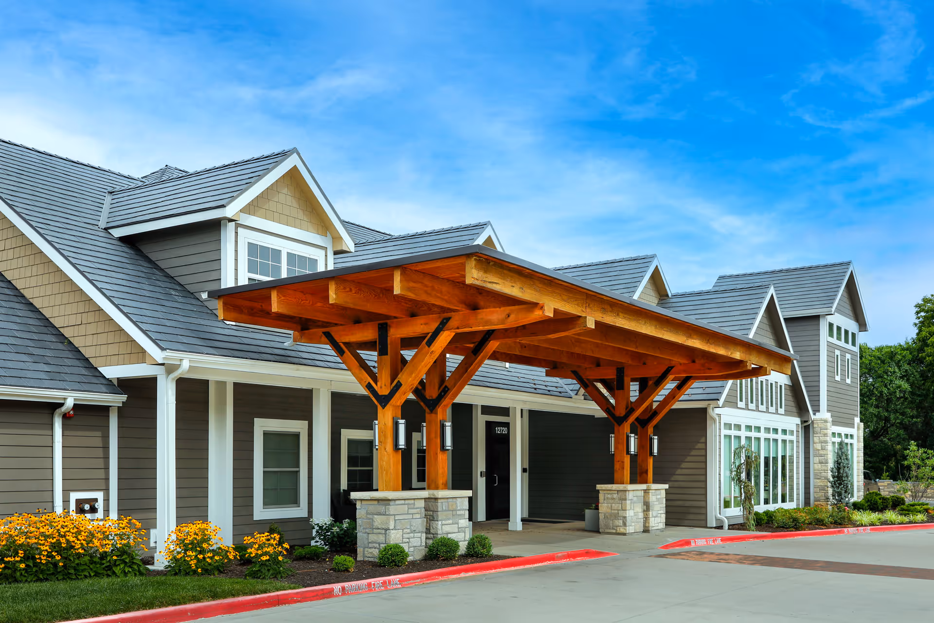 Exterior view of Homestead Estates Assisted Living of Leawood showing the building entrance with a large wooden canopy supported by stone pillars, surrounded by landscaping with yellow flowers and green shrubs under a blue sky.