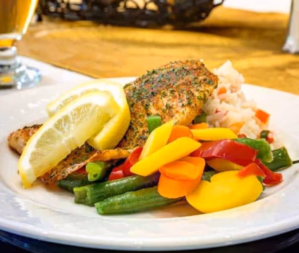 A plate of seasoned grilled fish garnished with lemon wedges, served with steamed green beans, sliced carrots, yellow and red bell peppers, and a side of rice with diced vegetables.