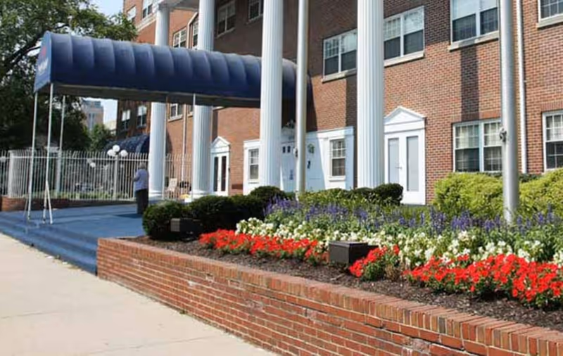 Entrance of a brick building with large white columns and a blue canopy over the steps. There is a flower bed with red, white, and purple flowers in front of the building, and a person standing near the entrance. The building has multiple windows and white doors.