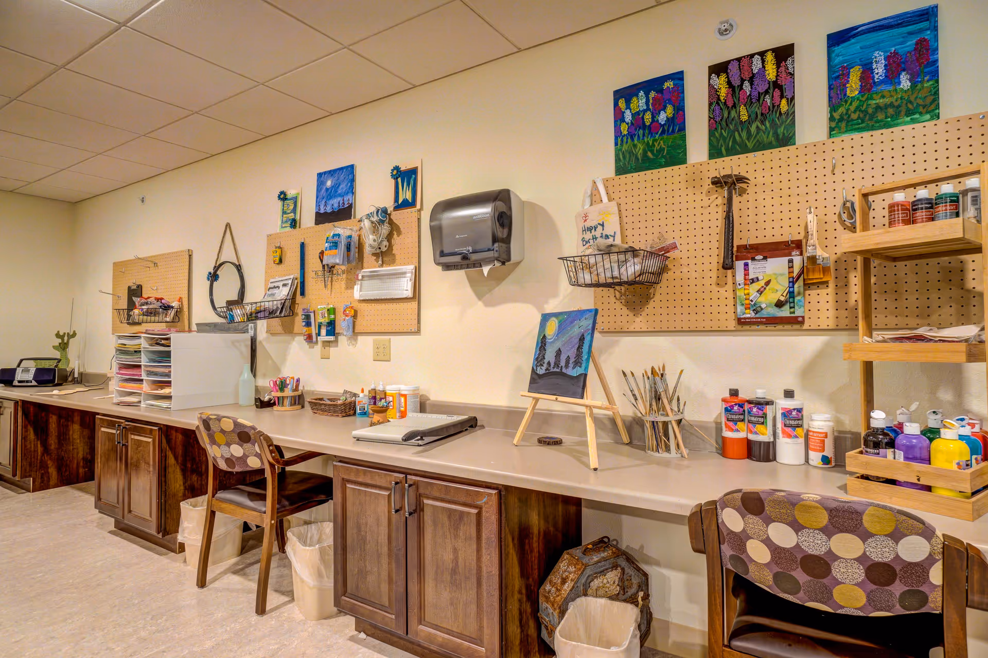 A well-organized art and craft room with a long countertop, wooden cabinets, and pegboards on the wall holding various art supplies and small paintings. There are two chairs with patterned upholstery, paint bottles, brushes in containers, and a small easel displaying a painting of a night scene with trees and a moon.