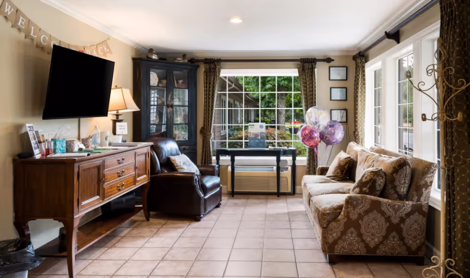 A cozy living room area with a patterned sofa on the right, a leather armchair next to a dark wooden cabinet with glass doors, and a wooden sideboard with a lamp and various items on top. A flat-screen TV is mounted on the wall above the sideboard. Large windows with curtains let in natural light and show greenery outside. There are balloons near the window and a 'Welcome Back' banner on the wall.