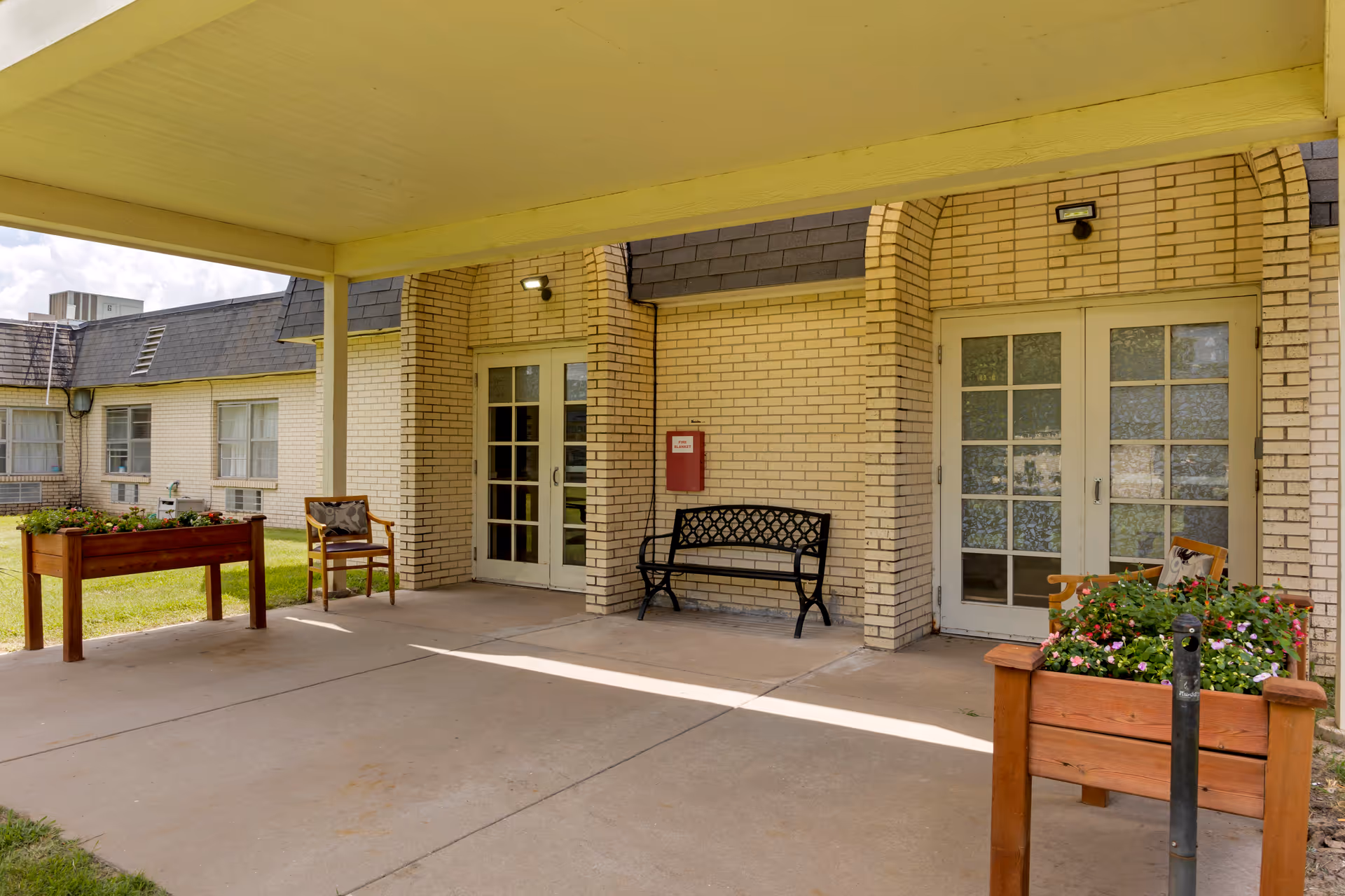 Covered entrance of a light-brick rehabilitation center showing double glass doors, benches, chairs, and wooden planter boxes.
