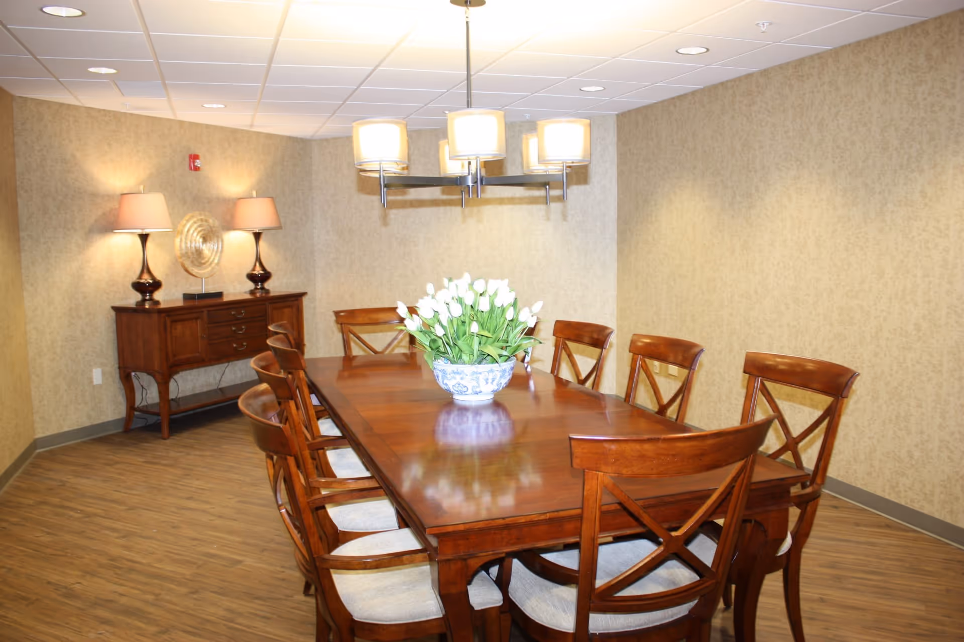 A dining room with a large wooden table surrounded by eight wooden chairs with cushioned seats. A decorative bowl with white flowers is placed in the center of the table. The room has beige patterned wallpaper, a wooden sideboard with two lamps and a decorative plate, and a modern chandelier hanging above the table. The floor is wood-patterned.