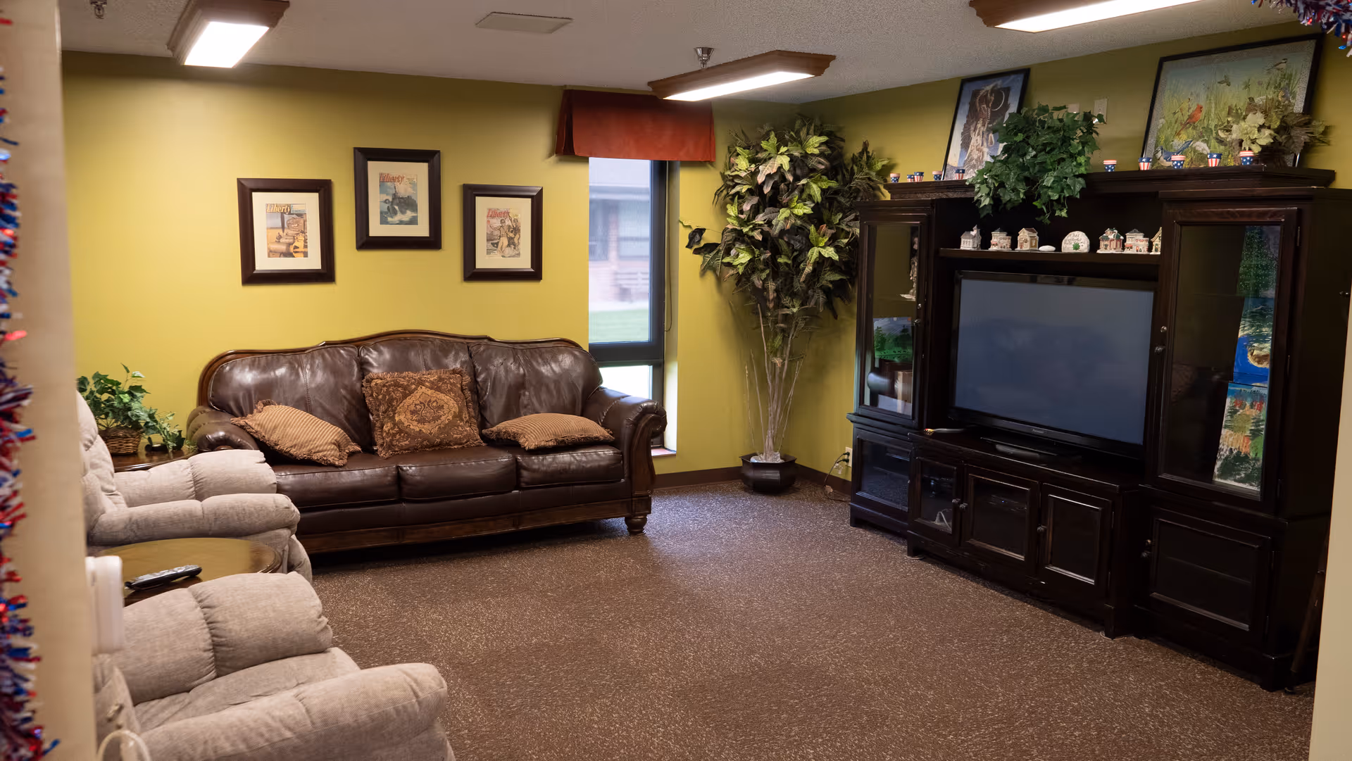 Communal living room with a leather sofa, recliners, large entertainment center with TV, and decorative plants and artwork on green walls.