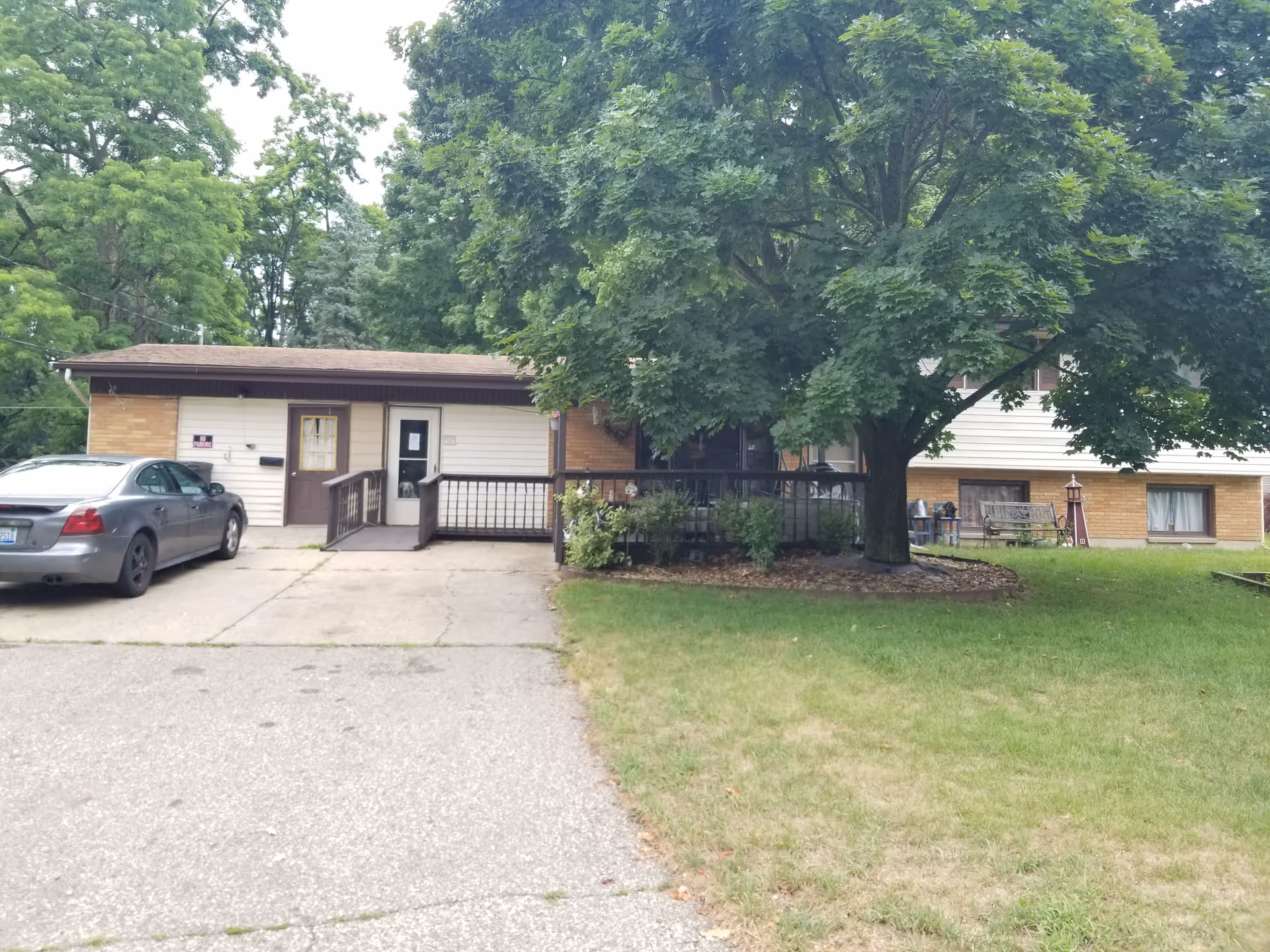 Exterior view of a single-story residential building with a brown roof and light-colored siding. There is a large tree with green leaves in front of the building, a small porch with a ramp, and a parked silver car on the driveway. The lawn is green with some patches of dry grass.