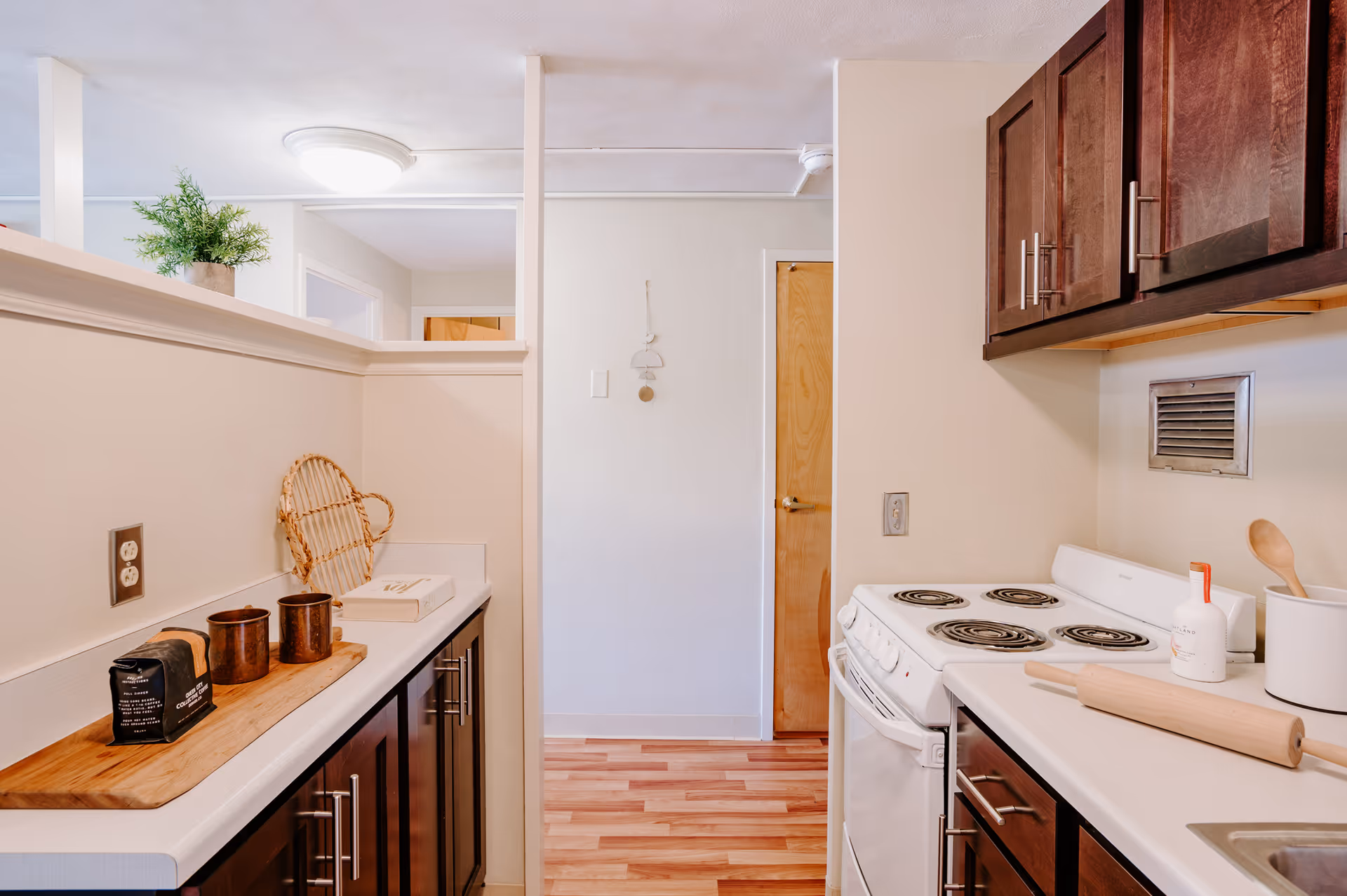 A small kitchen area with dark wooden cabinets, a white electric stove, a rolling pin, and kitchen utensils on the counter. There is a wooden cutting board with two brown cups and a black package on the left counter. The floor is wooden, and there is a door at the end of the kitchen space.