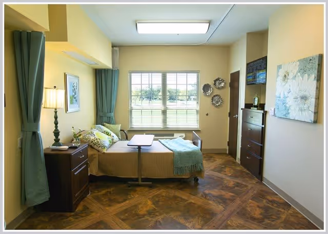 A well-lit bedroom in a senior living facility with a single bed positioned near a large window with green curtains. The bed has a brown bedspread, green and floral pillows, and a teal throw blanket. Next to the bed is a wooden nightstand with a lamp and a small plant. Across from the bed is a tall wooden dresser with a flat-screen TV mounted above it and decorative wall art. The room has wood-patterned flooring and light yellow walls.