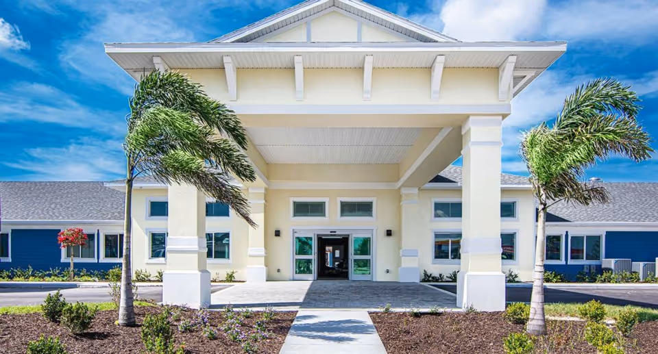 Front exterior view of Hampton Manor of Cape Coral building with a covered entrance, two palm trees, and a clear blue sky.