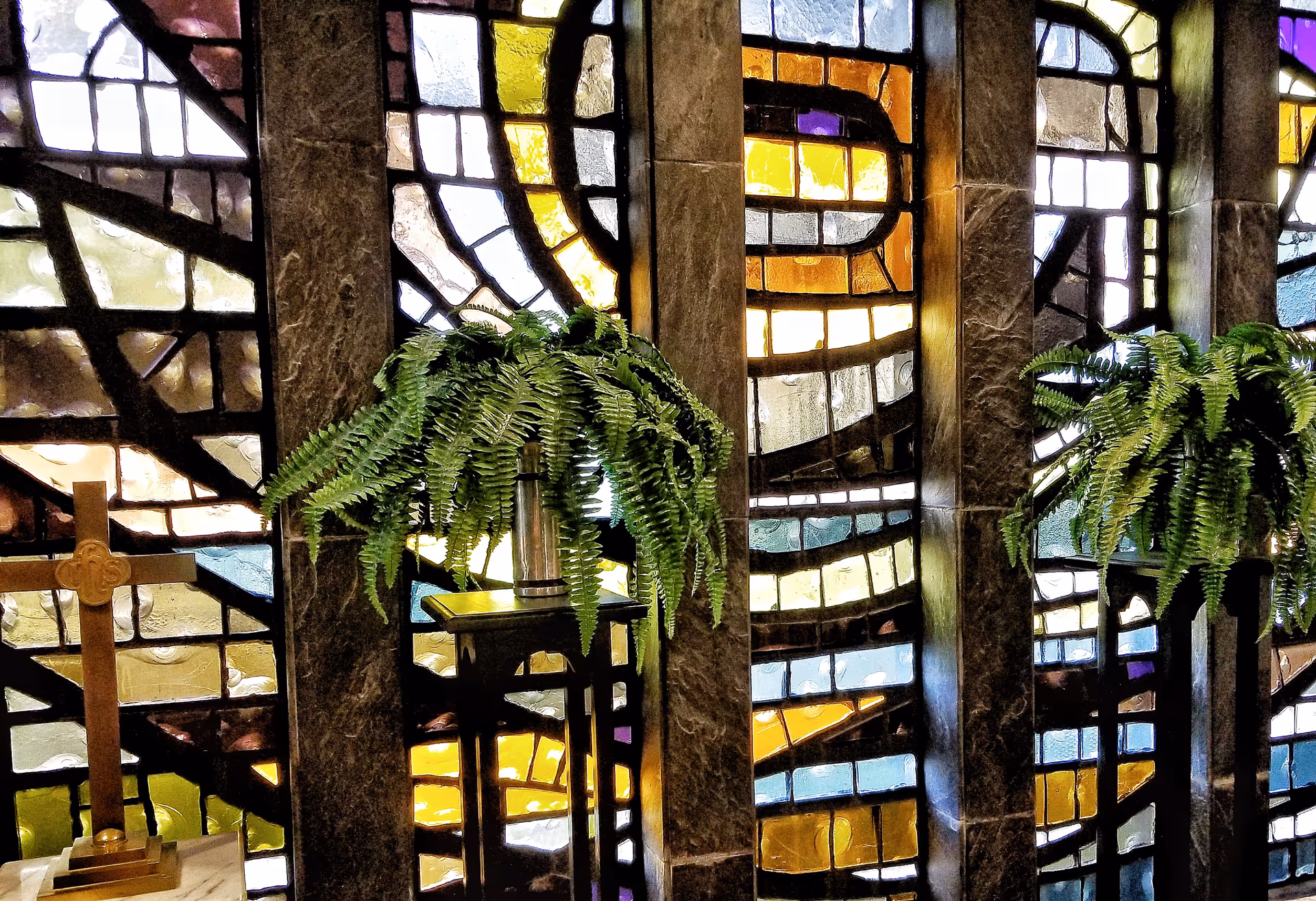 Interior view of a room with colorful stained glass windows featuring abstract patterns. Two green fern plants are placed on tall black stands in front of the windows, and a wooden cross is visible on the left side.