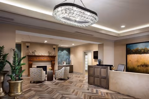 A spacious and elegantly decorated living area featuring a modern chandelier, two patterned armchairs facing a wooden fireplace mantel, a reception desk with a chair, a large potted plant, and a large landscape painting on the wall.