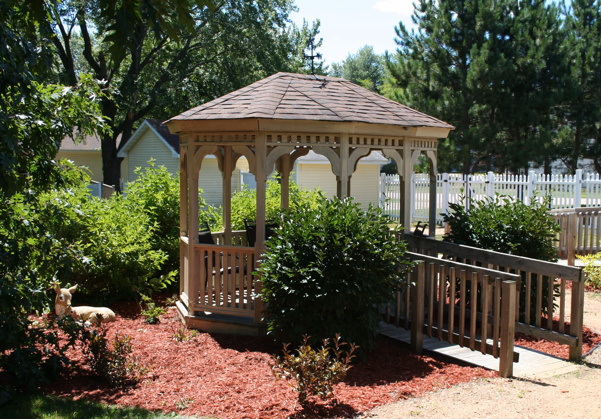 A wooden gazebo with a shingled roof surrounded by green bushes and red mulch in an outdoor garden area. There is a wooden ramp leading up to the gazebo, and a white picket fence and trees are visible in the background.