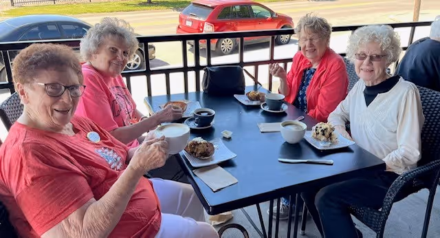 Four elderly women sitting around a black outdoor table enjoying coffee and pastries on a patio with a street and parked cars visible in the background.