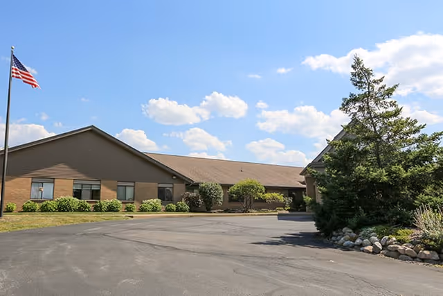 Exterior view of a single-story brick building with a sloped roof under a blue sky with scattered clouds. There is an American flag on a flagpole to the left and landscaped bushes and trees around the building. The paved driveway curves in front of the building entrance.