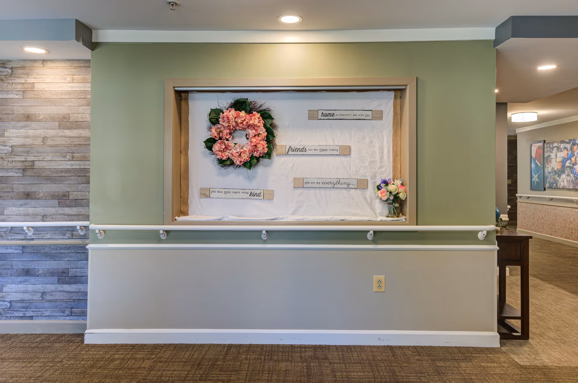 A hallway wall display in a senior living facility featuring a pink floral wreath and inspirational wooden plaques with messages about home, friends, and kindness. The wall is painted green and beige with a handrail running along it. To the left is a section of wall with gray wood paneling, and to the right is a glimpse of another hallway with artwork on the walls.
