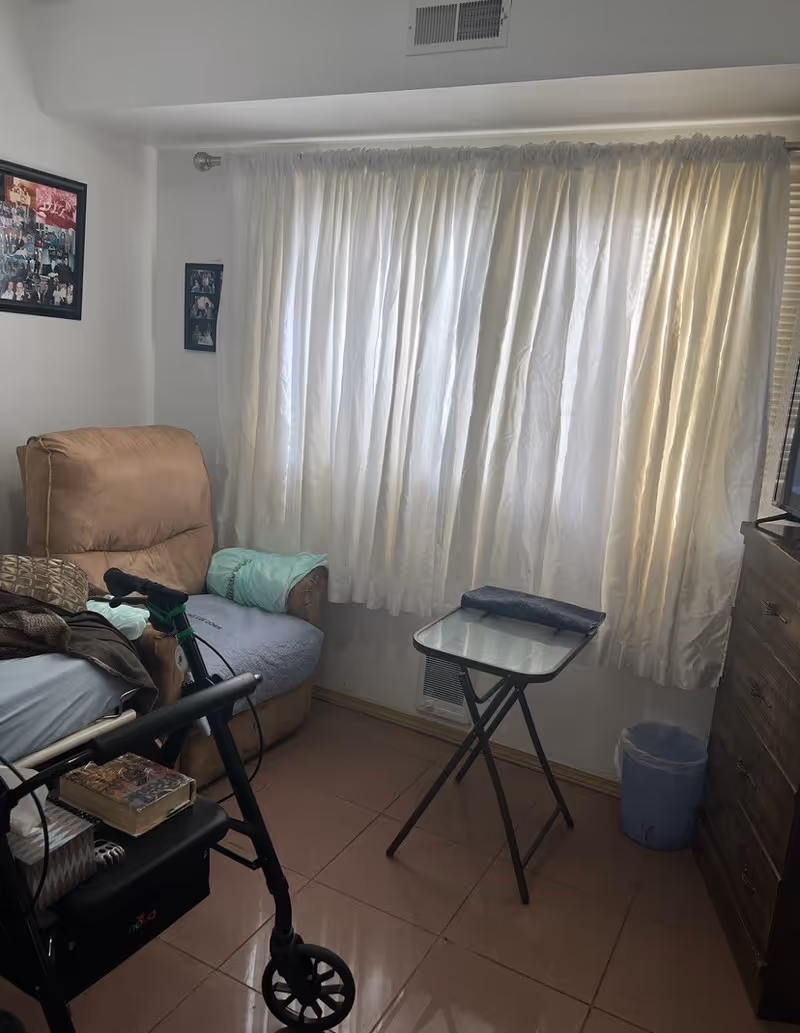 A small room with a beige recliner chair covered partially with a light blue blanket, a walker with a book and tissue box on its seat, a small glass-top folding table, a wooden dresser, a blue trash bin, and white curtains covering a window. There are framed photos on the white walls.