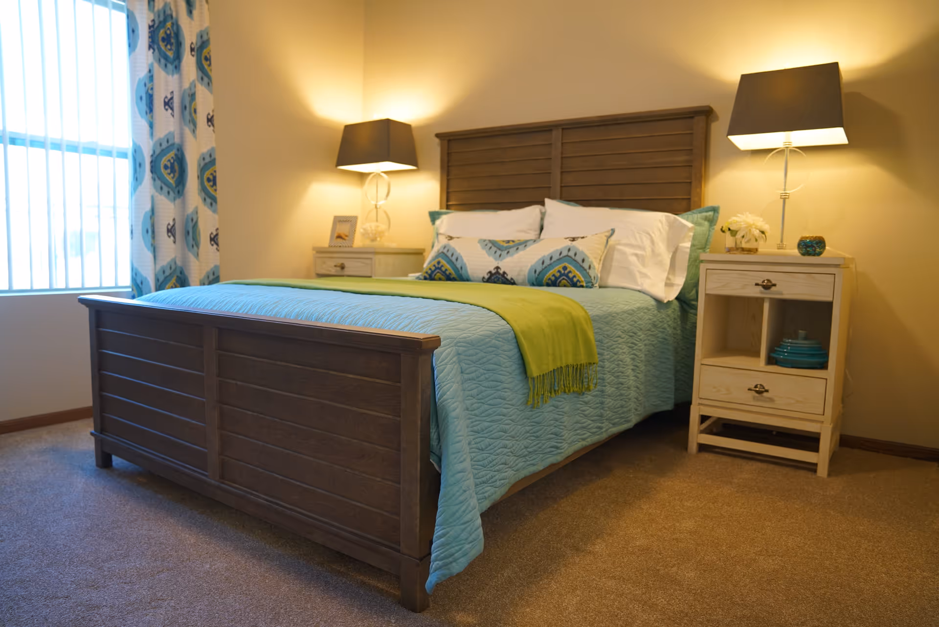 A cozy bedroom with a wooden bed frame, a blue quilted bedspread, decorative pillows, and a green throw blanket. There are two matching nightstands on either side of the bed, each with a lamp and decorative items. A window with patterned curtains is visible on the left side, and the room is softly lit with warm lighting.