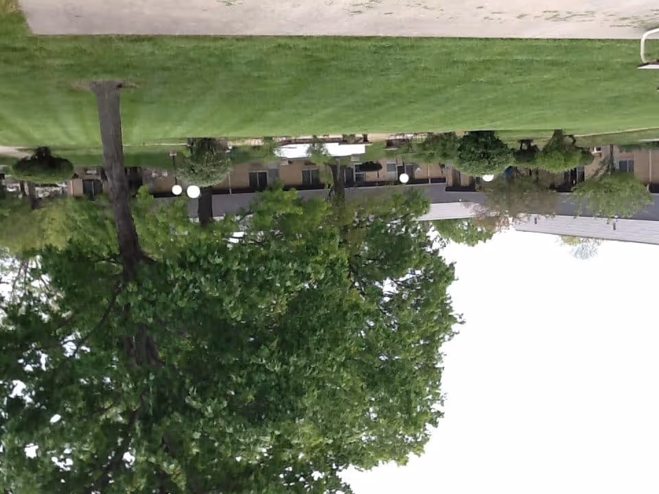 View of a senior living facility building with a large green lawn and several trees in the foreground. The building has multiple windows and a gray roof, with outdoor lighting fixtures visible along a walkway.