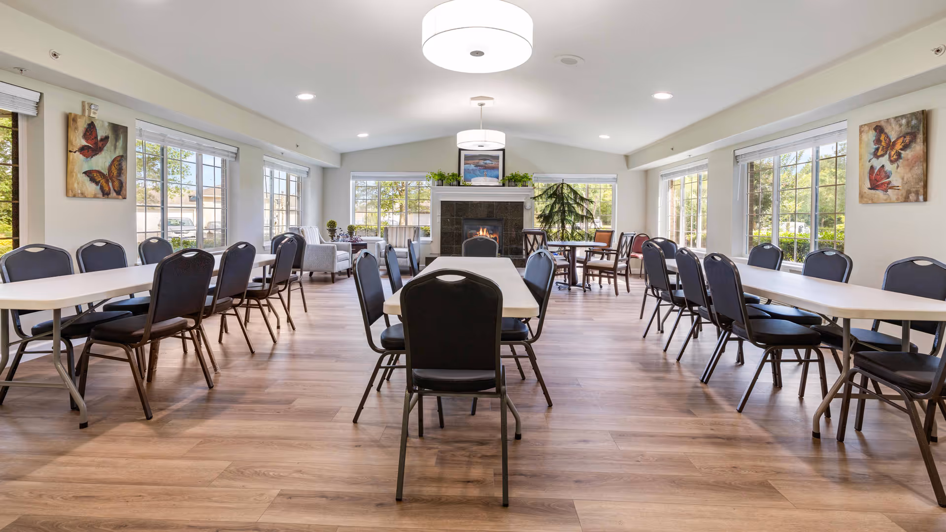 A bright and spacious dining room with multiple rectangular tables and black chairs arranged neatly. Large windows on three sides allow natural light to fill the room. There is a fireplace at the far end with a painting and plants on the mantel. Butterfly artwork hangs on the walls, and the floor is a light wood laminate.