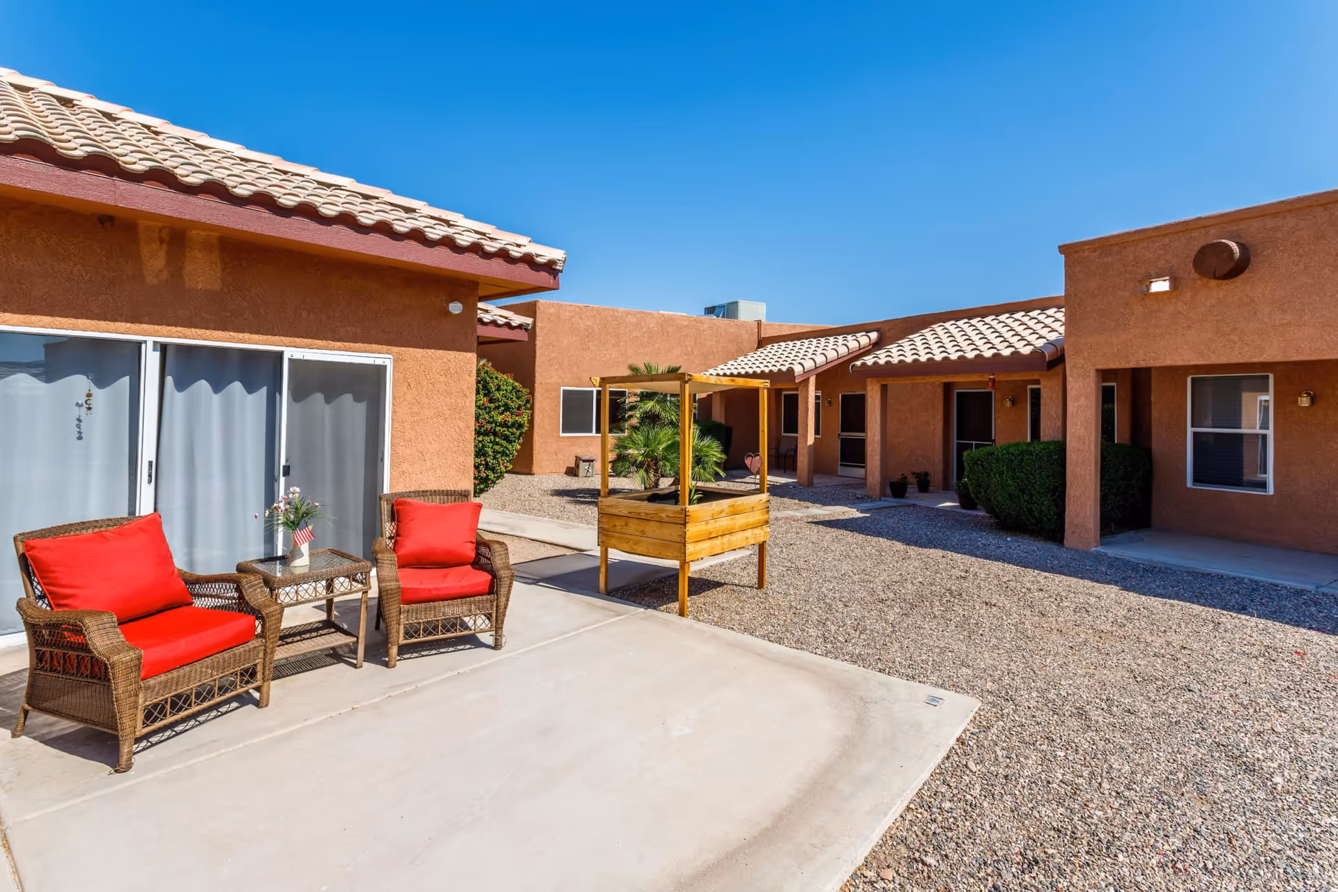 Outdoor courtyard of a terracotta stucco assisted living building with two wicker chairs with red cushions, a small table, a raised wooden planter and gravel walkways under a clear blue sky.