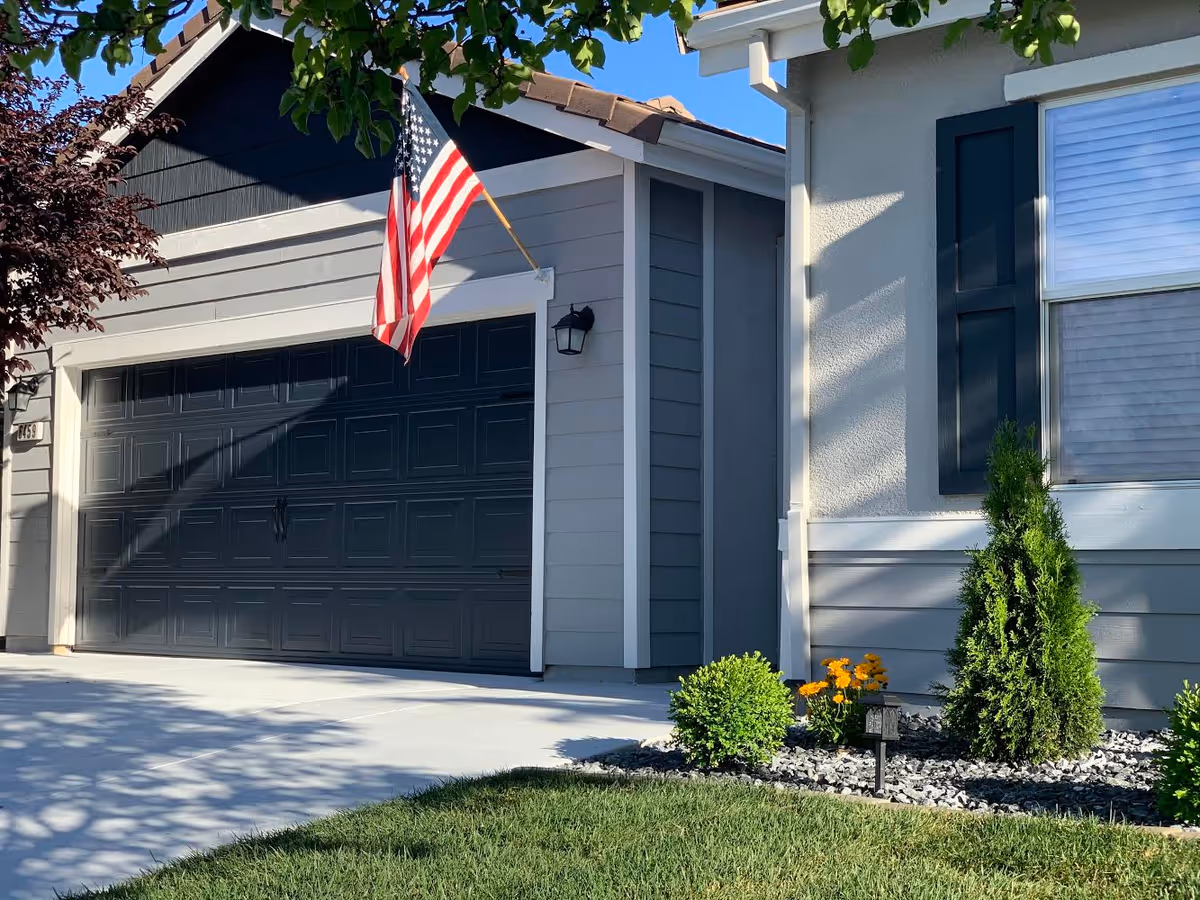 Exterior view of a residential building with a closed dark gray garage door, an American flag mounted above the garage, a small landscaped area with green shrubs and yellow flowers, and part of a window with dark shutters.