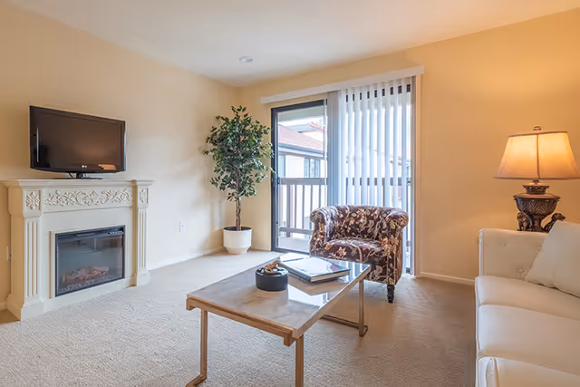 A cozy living room with a white sofa, a floral patterned armchair, a wooden coffee table with books and a decorative item, a potted plant in the corner, a wall-mounted TV above an ornate electric fireplace, and a sliding glass door with vertical blinds leading to a balcony.