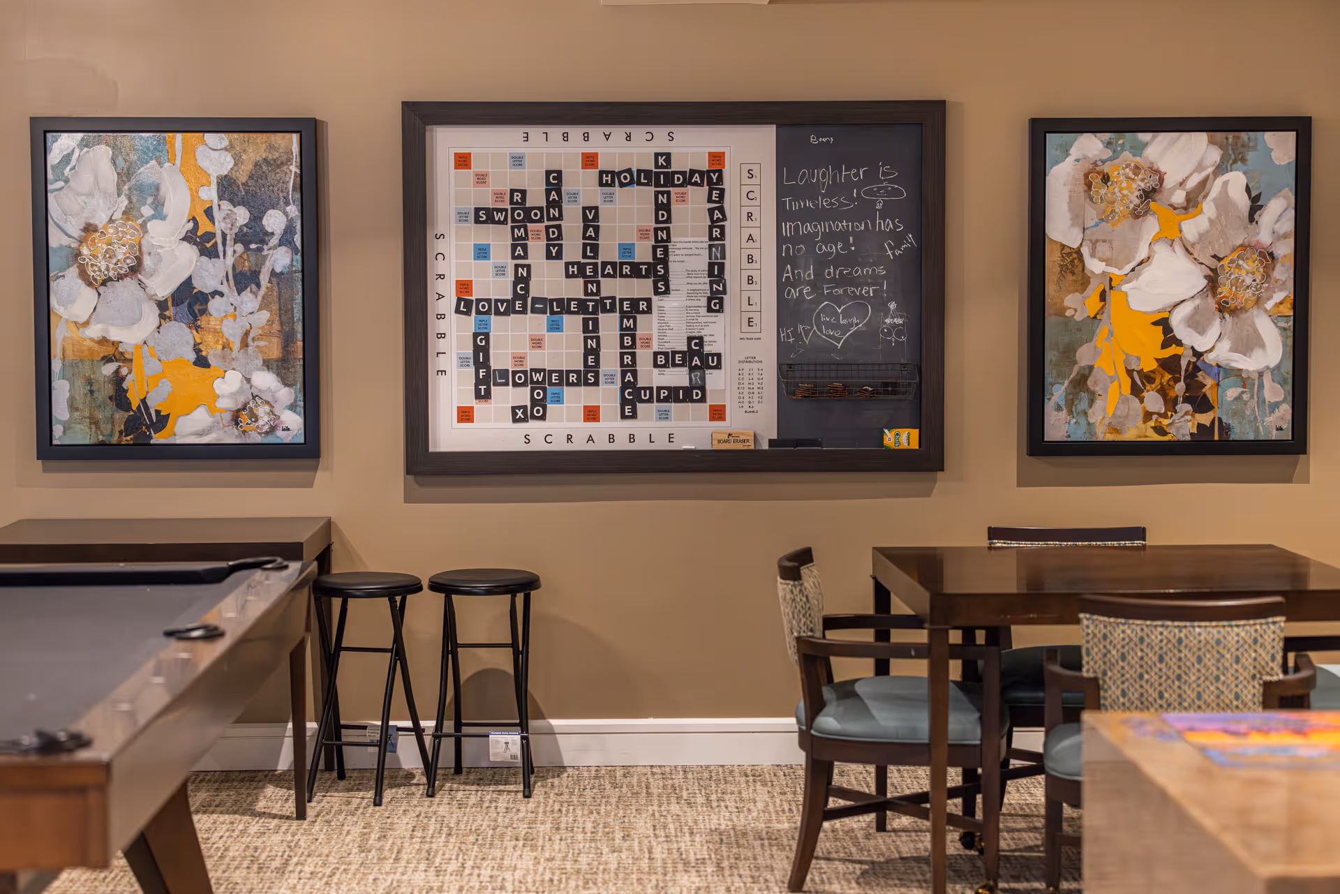 A cozy game room with a pool table on the left, two black bar stools against the wall, and a wooden table with four chairs on the right. On the wall, there is a large framed Scrabble board with words arranged on it and a chalkboard section with handwritten inspirational quotes. Two framed floral paintings flank the Scrabble board on either side. The room has beige walls and carpeted flooring.