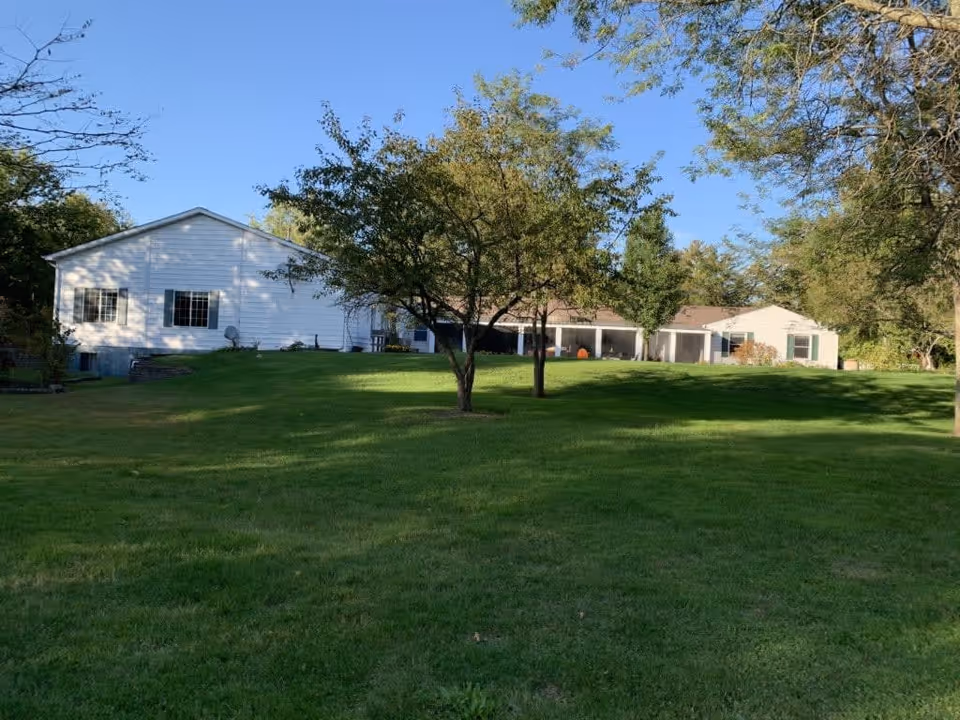 A single-story white building with multiple windows and a covered porch, surrounded by a large green lawn with several trees under a clear blue sky.