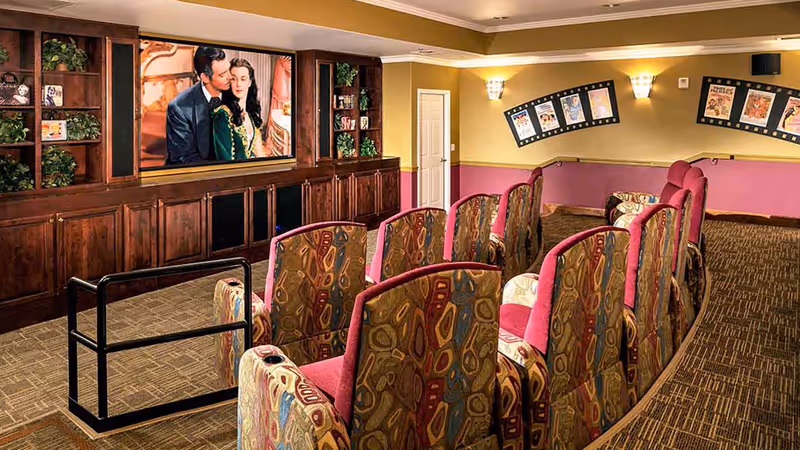 A cozy movie theater room with patterned upholstered chairs arranged in rows facing a large screen displaying a classic movie scene. The room has warm lighting, wood cabinetry with shelves holding decorative items, and movie posters on the walls.