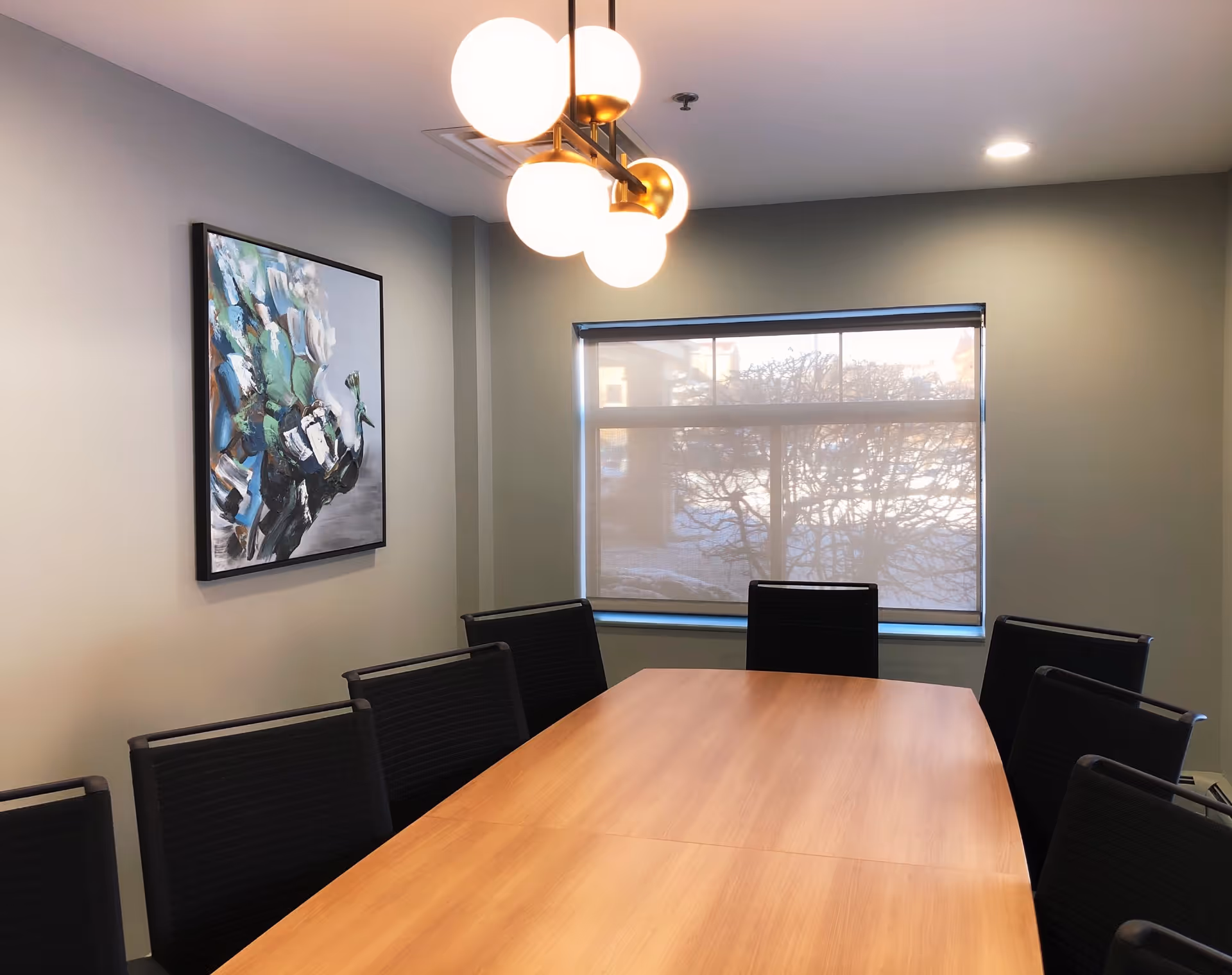 A modern conference room with a wooden table surrounded by black chairs. There is a large window with a roller shade partially down, and a contemporary light fixture with round white bulbs hanging from the ceiling. A colorful abstract painting is mounted on the gray wall.