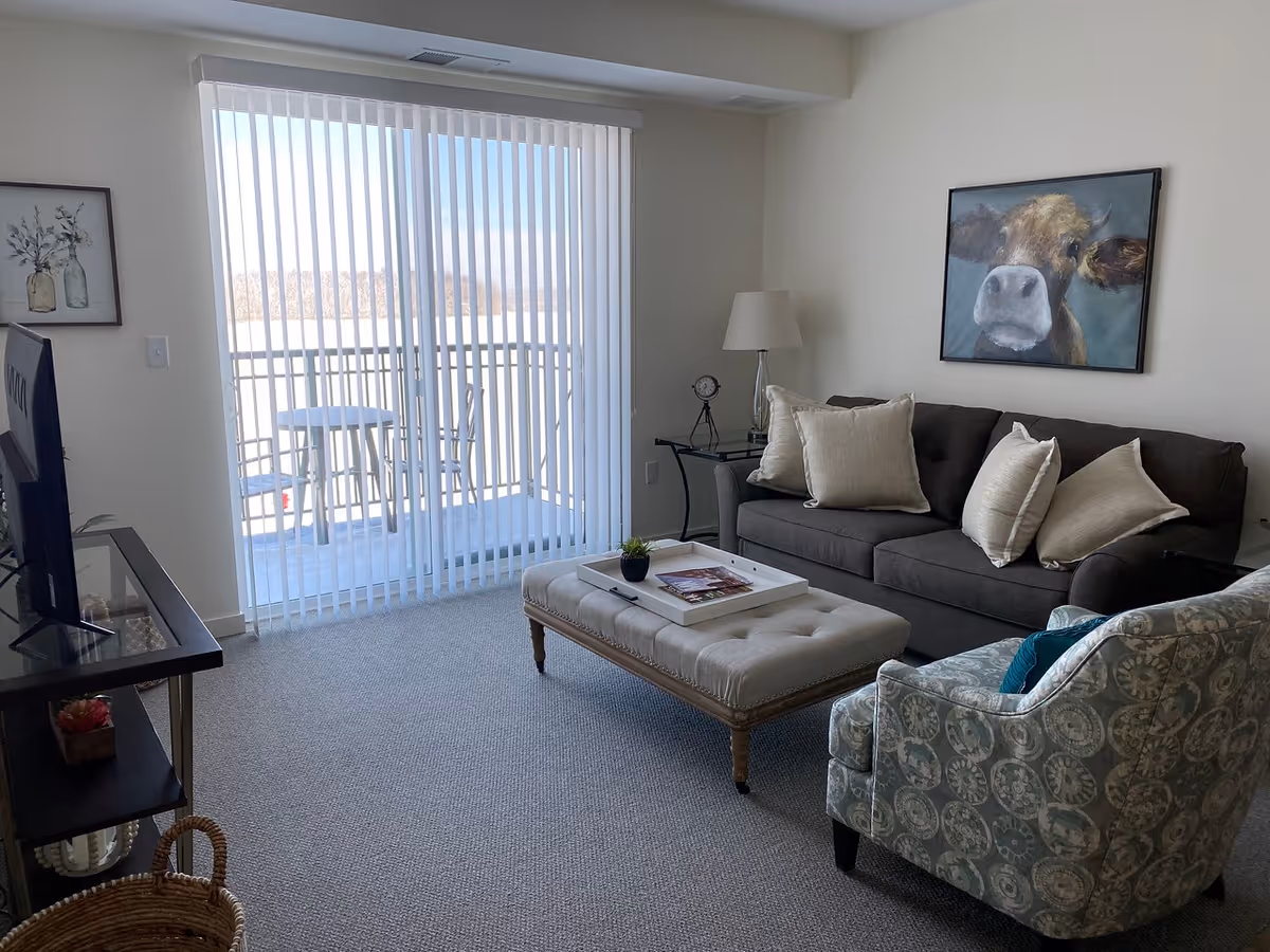 Bright living room with a dark sofa, patterned armchair, tufted ottoman, TV stand, and sliding glass door opening to a balcony.