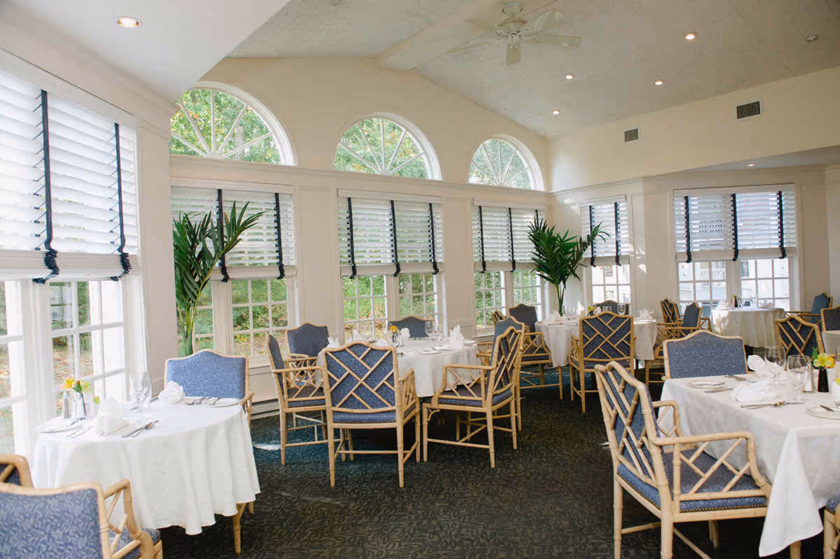 A bright sunlit dining room with round white-clothed tables, blue-cushioned chairs, large arched windows, and potted plants.