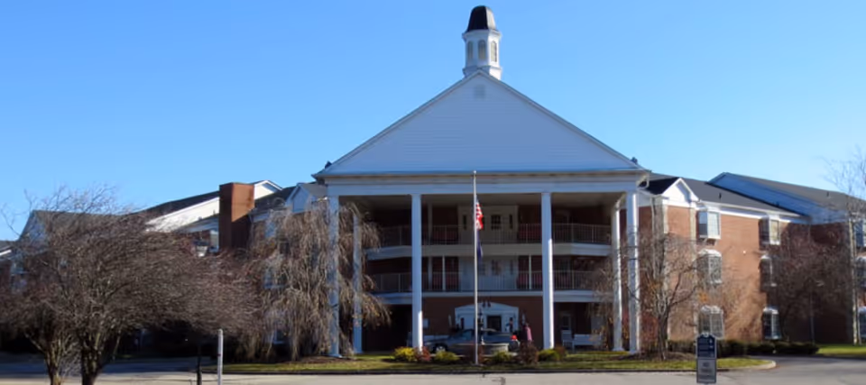 Front exterior view of a large senior living facility named Colonial Gardens with a white triangular pediment, tall white columns, multiple balconies, and an American flag in front. The building is made of red brick with white trim and surrounded by leafless trees and a parking area.