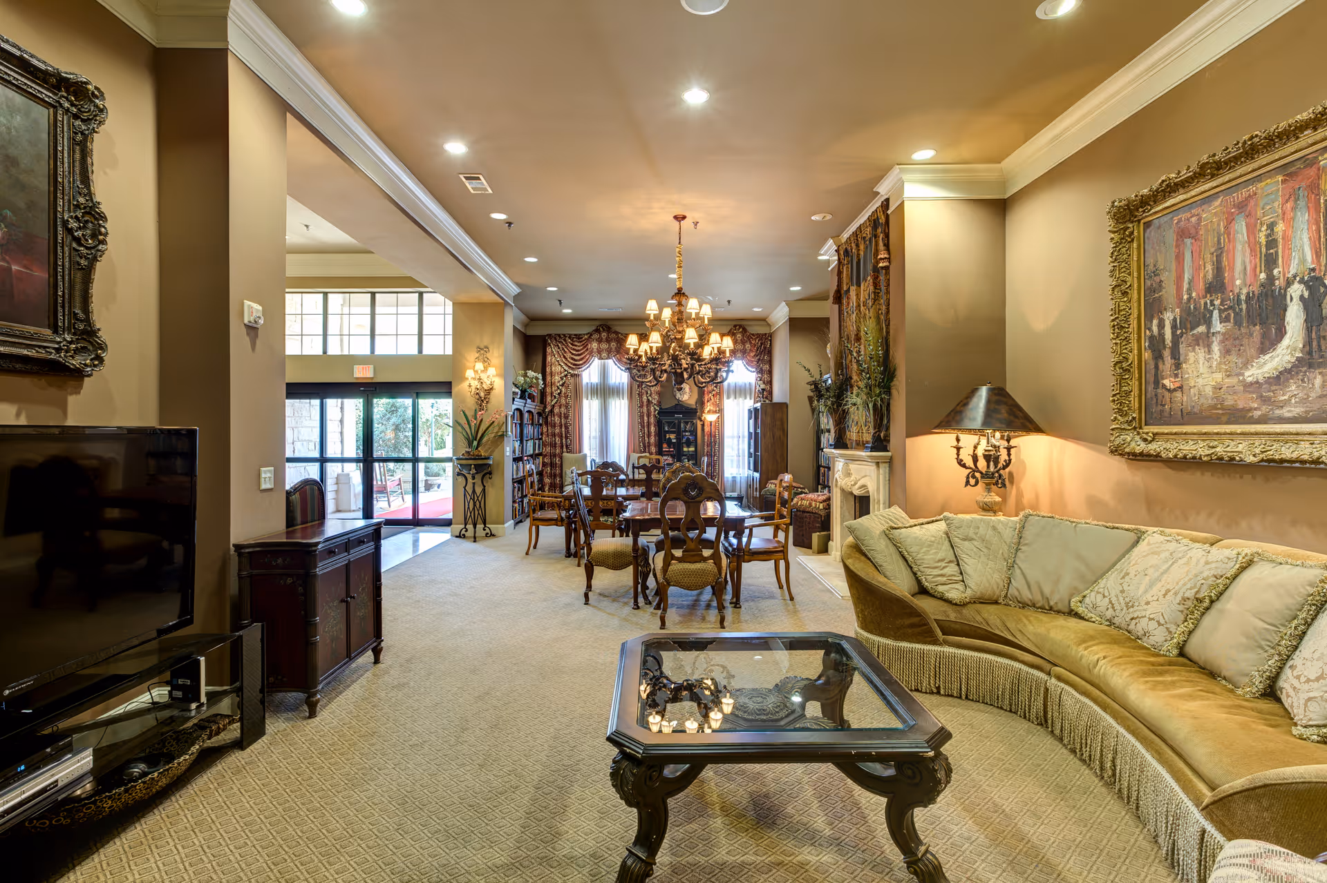 Interior view of a senior living facility lounge and dining area featuring a curved beige sofa with patterned cushions, a glass-top coffee table, ornate framed paintings on the walls, a chandelier above a wooden dining table with chairs, and large windows with draped curtains allowing natural light.