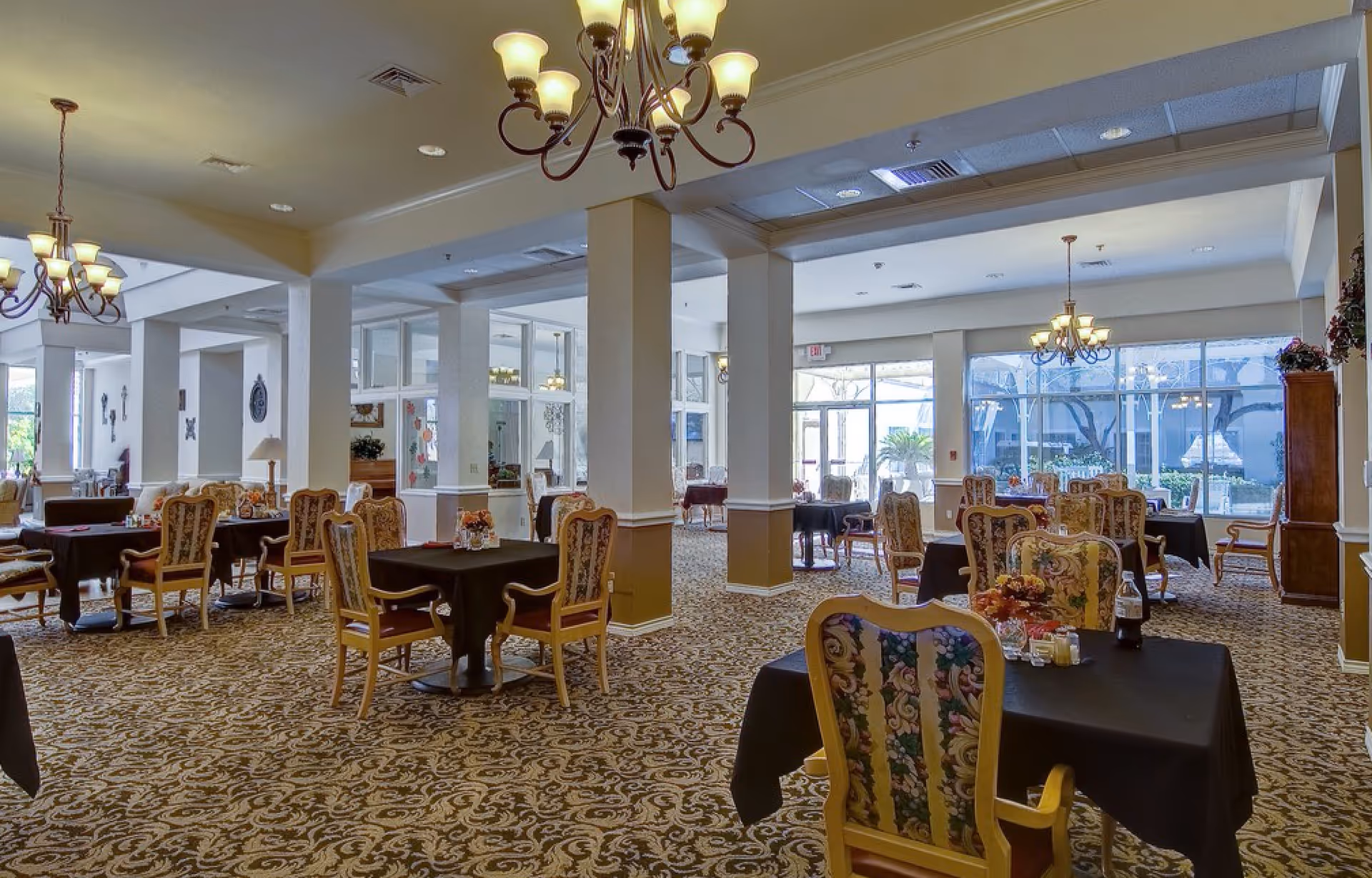 Spacious dining room with multiple tables and upholstered chairs, chandeliers, and large windows overlooking an outdoor courtyard.