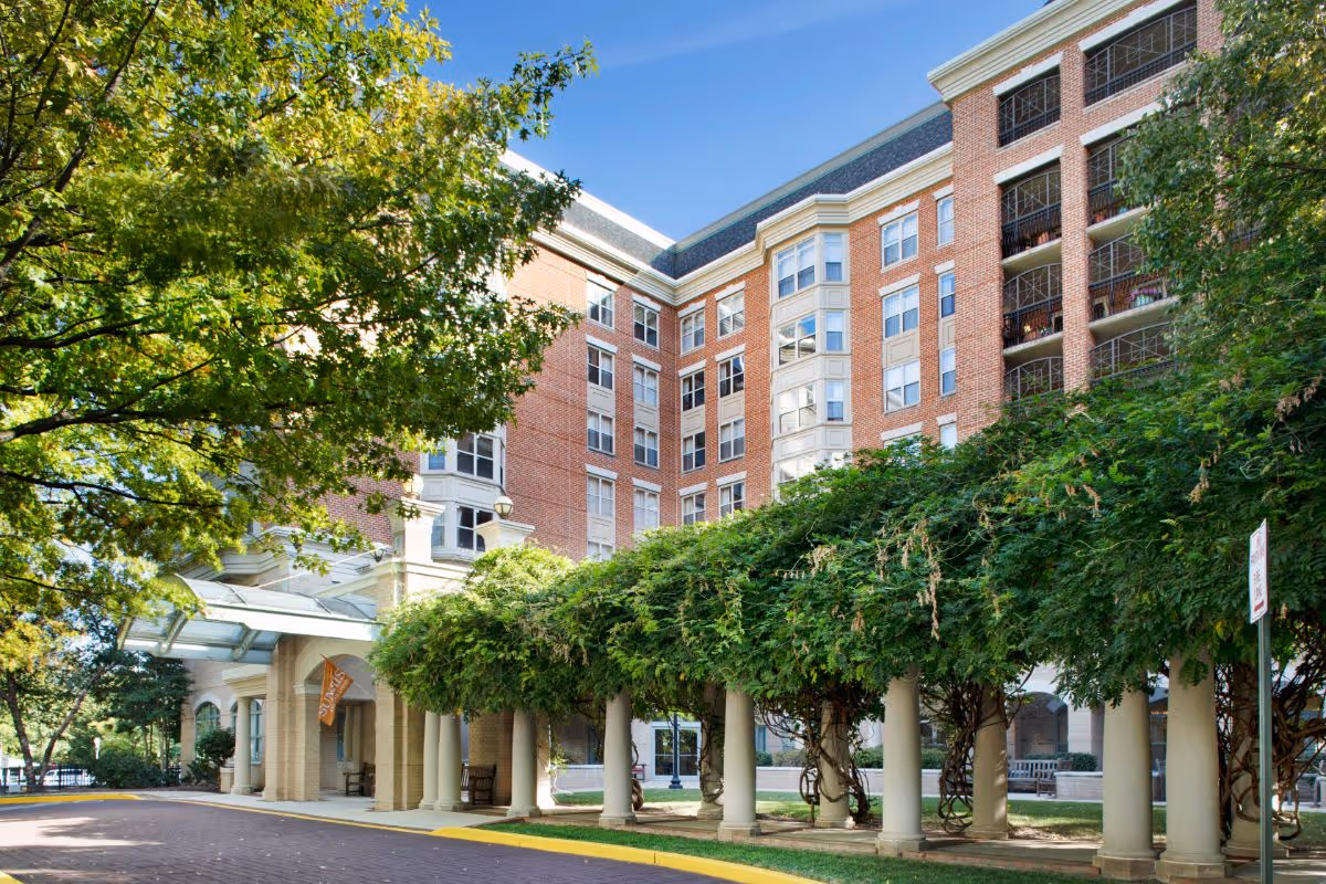 Exterior view of a multi-story brick building with large windows and balconies, surrounded by trees and greenery. A covered walkway with columns and lush green vines leads to the entrance, with benches placed along the path. The sky is clear and blue.