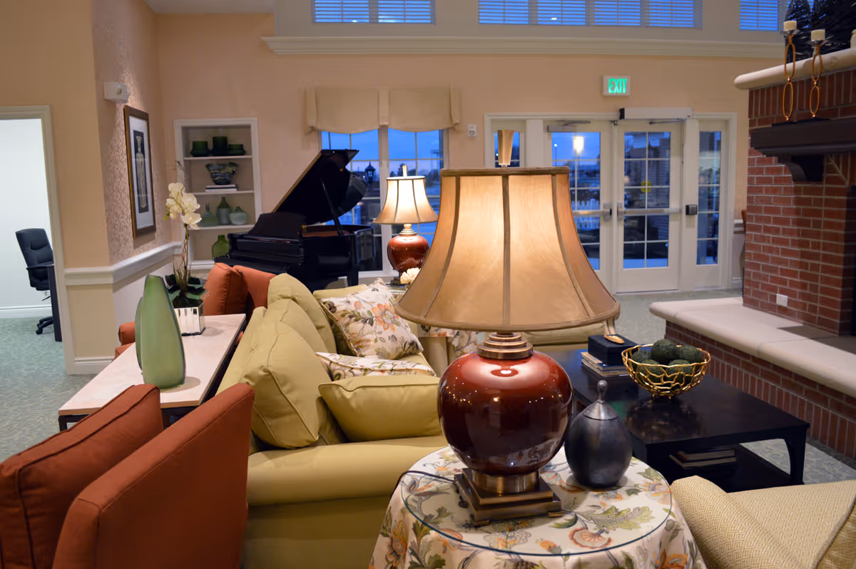 Cozy common living room with upholstered sofas, table lamps, a grand piano, and a brick fireplace near glass exit doors.