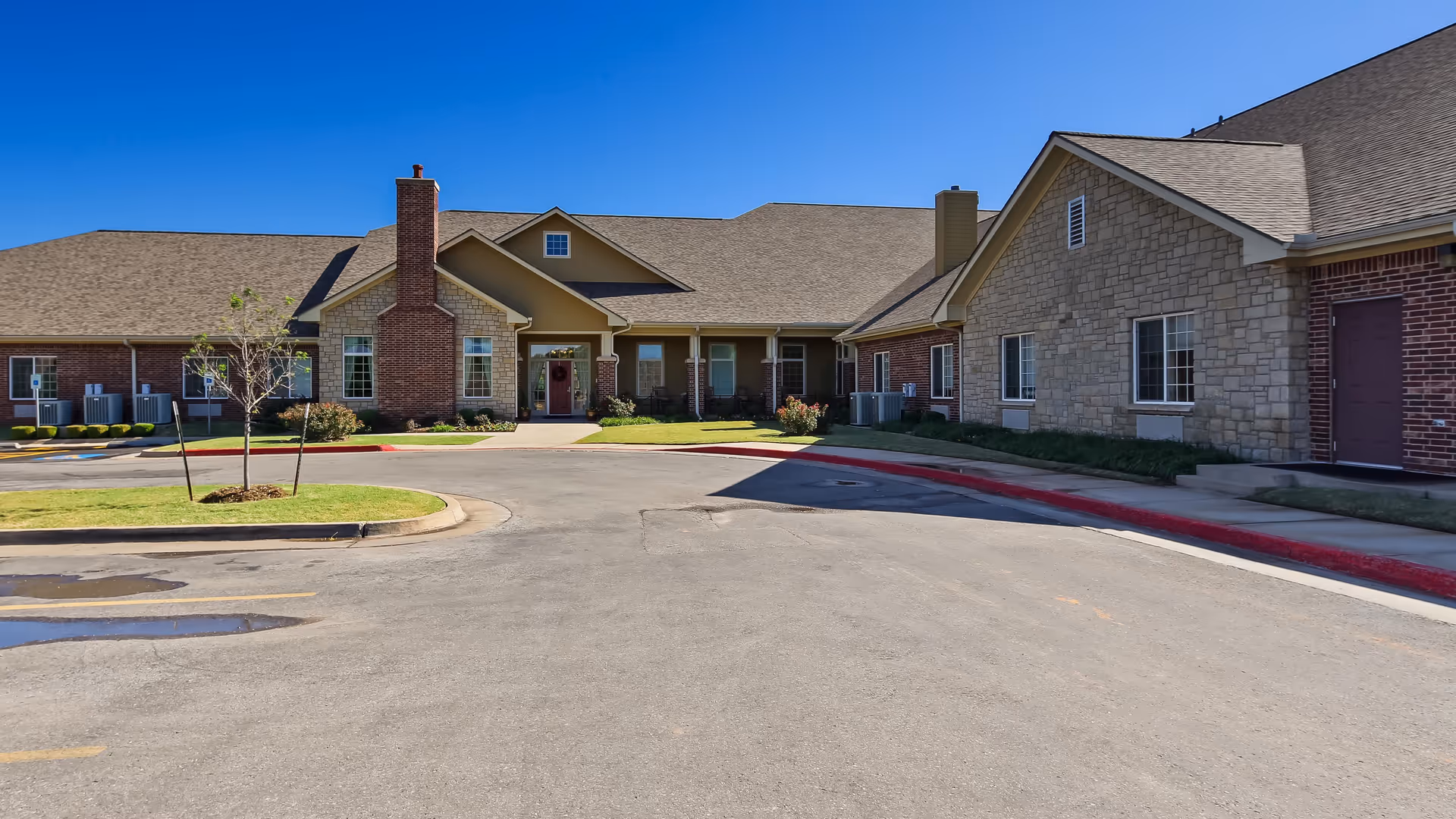 Exterior view of a single-story senior living facility building with brick and stone facade, a driveway in front, small landscaped areas with young trees, and a clear blue sky overhead.