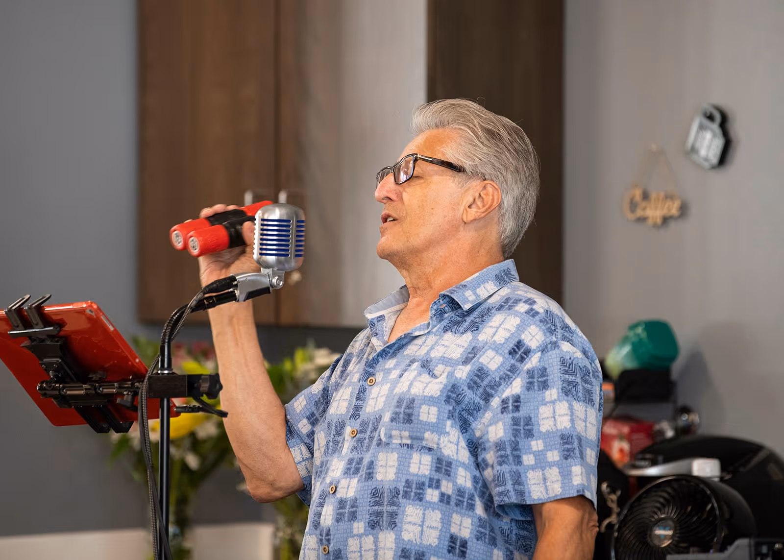 An elderly man wearing glasses and a blue patterned shirt is singing into a vintage-style microphone while holding a red shaker instrument in his hand. He is standing indoors with a kitchen area visible in the background, including cabinets and some kitchen items.