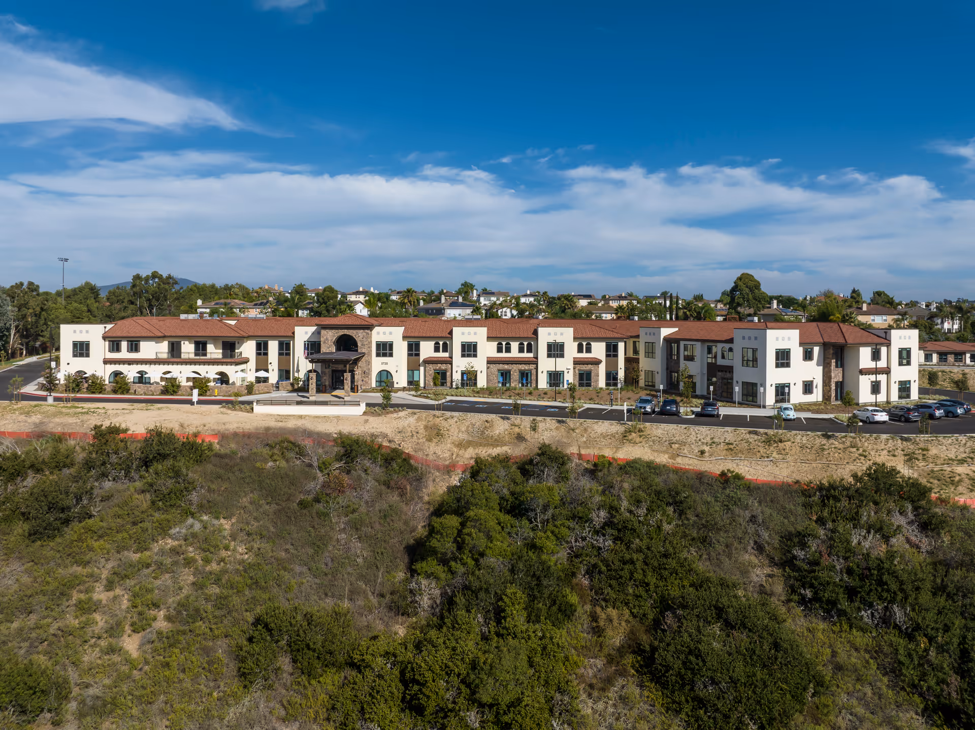 Wide exterior view of Westmont of Carmel Valley, a large two-story senior living facility with a beige facade and red tile roof, surrounded by greenery and a parking lot with several cars under a partly cloudy blue sky.