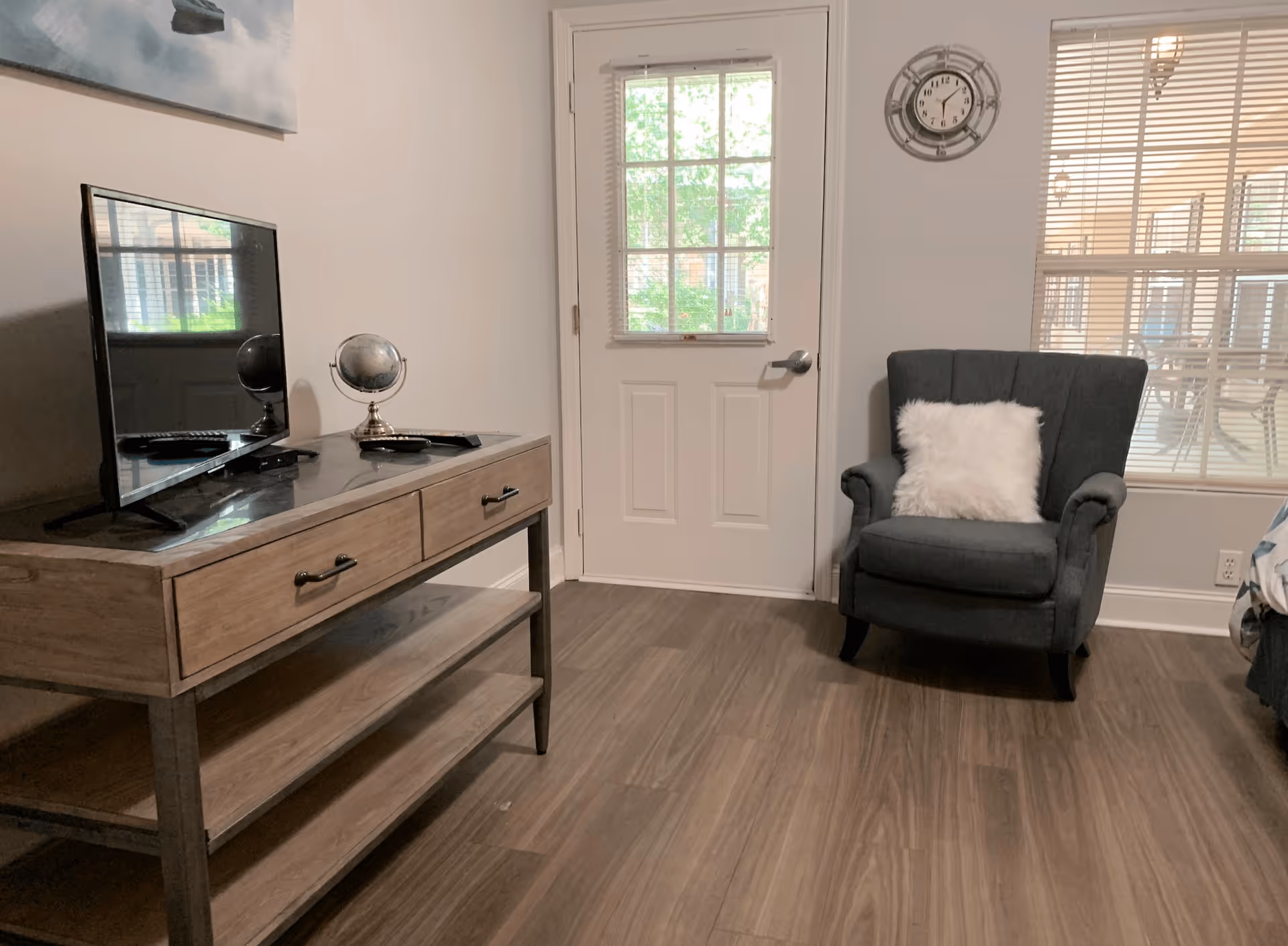 A cozy interior room with a wooden floor, a wooden console table holding a flat-screen TV and a small globe, a dark gray armchair with a white fluffy pillow, a white door with a window, a wall clock, and a window with blinds.
