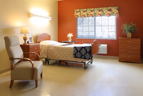 A well-lit senior living bedroom with a single bed covered in beige bedding and a dark patterned throw. There is a wooden nightstand with a lamp and a framed photo next to the bed. A comfortable upholstered armchair with wooden armrests is positioned near the bed. The room has a large window with floral valance curtains, a wooden dresser with a potted plant, and a wall-mounted light fixture above the bed.