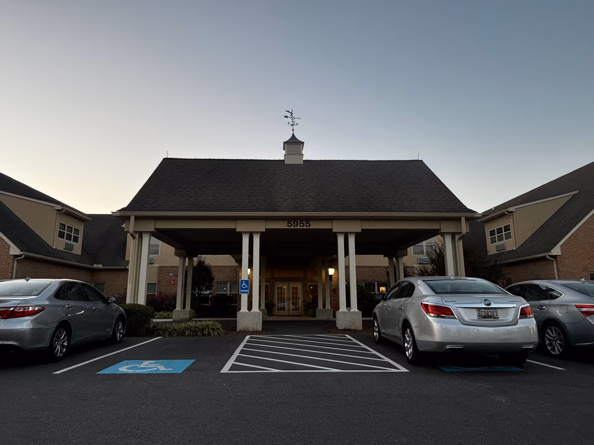 Front entrance of a retirement community building with a covered drop-off area supported by columns, a weather vane on the roof, and two cars parked in front including a handicapped parking space.