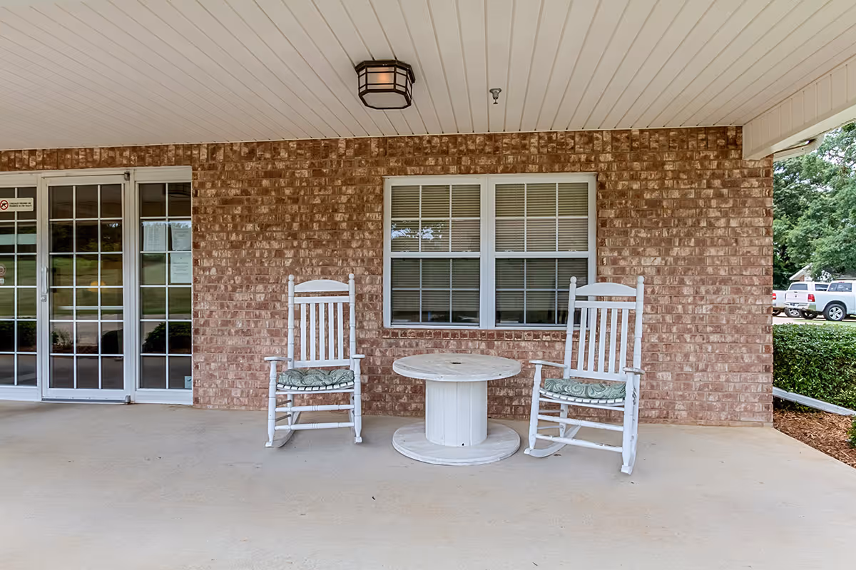 Covered front porch with two white rocking chairs and a round table in front of a brick wall and glass entrance doors.